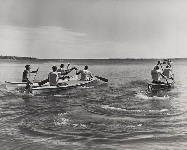 Rowers in three canoes paddle with oars in their hands across a lake in the middle ground, with the horizon and sky in the background.